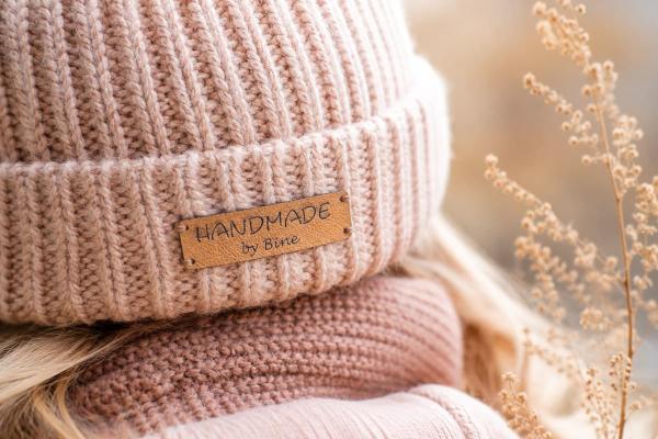 Etichetta rettangolare in similpelle beige "Handmade by Bine" su berretto rosa a maglia.